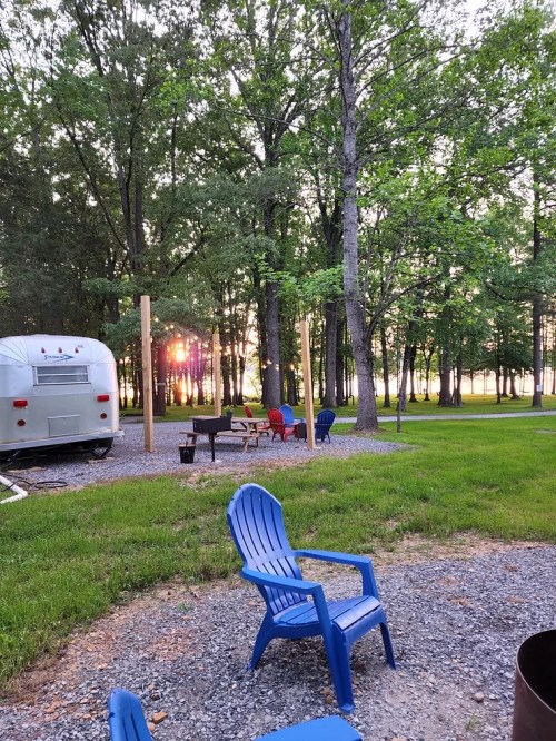 A serene campsite with blue chairs, a fire pit, and an Airstream trailer, surrounded by trees at sunset.