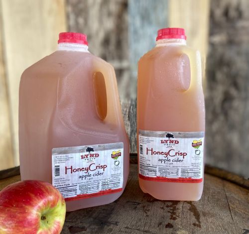 Two jugs of HoneyCrisp apple cider sit on a wooden surface, next to a fresh red apple.