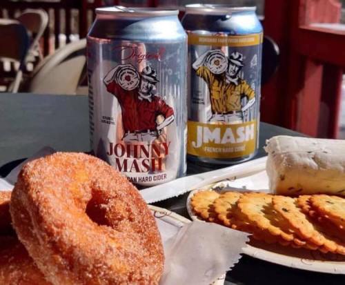 Two cans of hard cider labeled "Johnny Mash" and "J.Mash" next to a plate of donuts and crackers on a table.