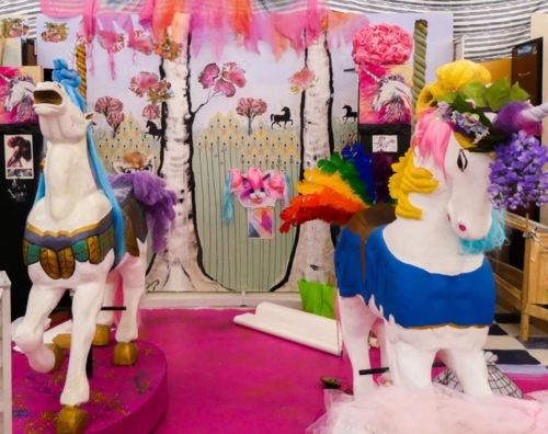 Colorful unicorn sculptures with vibrant decorations in a whimsical setting, featuring a backdrop of trees and flowers.