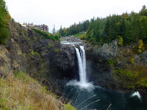 A waterfall cascades down rocky cliffs, surrounded by lush greenery and a distant building on the hillside.