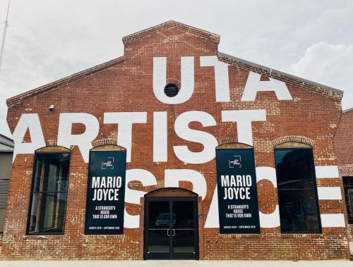 Exterior of a brick building with large text reading "UTA ARTIST SPACE" and banners for "MARIO JOYCE" exhibition.