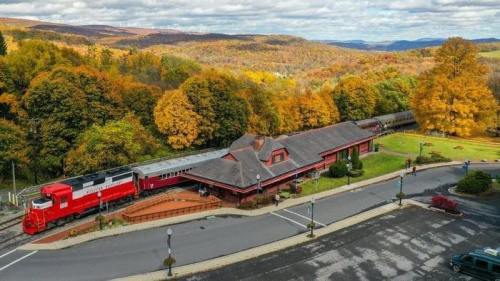 A red train at a scenic station surrounded by colorful autumn trees and rolling hills under a cloudy sky.