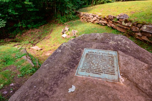 A plaque on a large rock in a grassy area, surrounded by trees and stone walls, commemorating a historical site.
