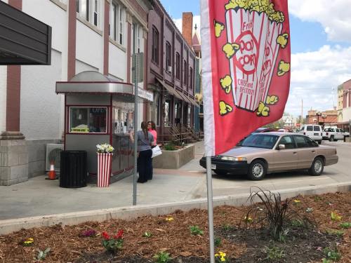 A popcorn stand with a red flag, flowers in the foreground, and a car parked nearby in a small town setting.