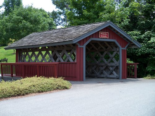 A red covered bridge with a wooden structure, labeled "Whisky Creek," surrounded by green trees and grass.