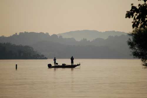 Two fishermen stand on a small boat in a calm lake at dawn, surrounded by misty hills and soft light.