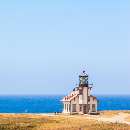 A white lighthouse with a glass lantern sits on a grassy shore overlooking a blue ocean under a clear sky.