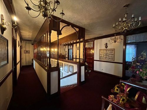 A well-lit hallway with wooden railings, chandeliers, and decorative walls, leading to a staircase.