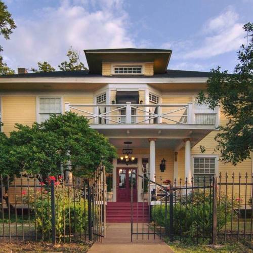 A charming yellow two-story house with a balcony, surrounded by greenery and a decorative iron fence.