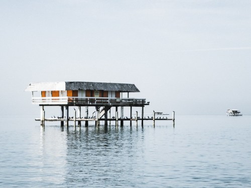 A wooden stilt house stands over calm water, reflecting the serene sky in a tranquil coastal setting.