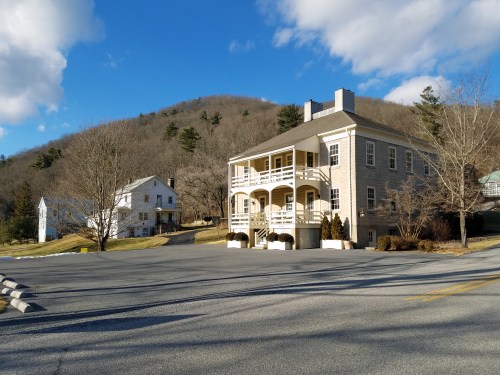 A two-story building with balconies, surrounded by trees and hills under a blue sky with clouds.