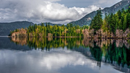 A serene lake surrounded by lush green trees and mountains, reflecting clouds and scenery on the water's surface.
