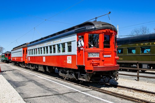A vintage red train car at a station, with a conductor waving from the window on a sunny day.