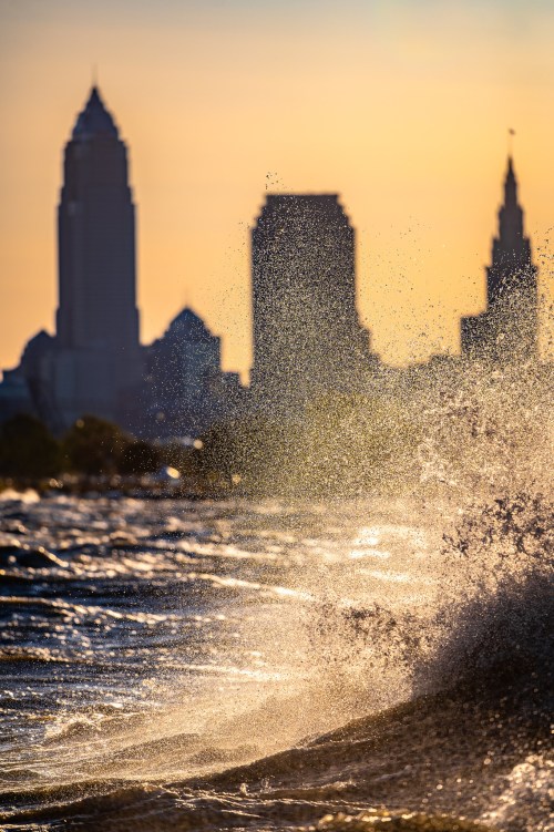 Silhouette of city skyline at sunset with waves splashing in the foreground, creating a misty effect.