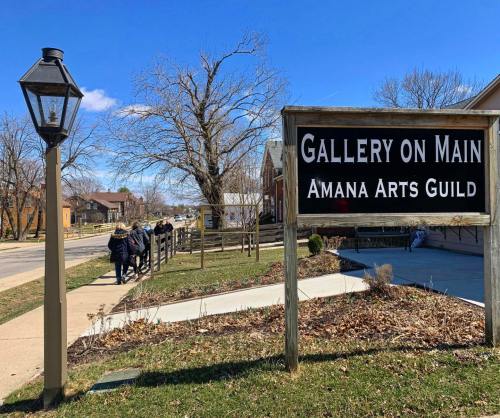 Sign for "Gallery on Main" with people walking by in a sunny, tree-lined street.