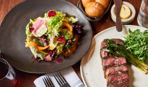 A plate with sliced steak and grilled greens, alongside a fresh salad and rolls on a wooden table.
