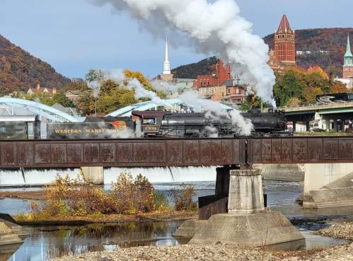 A steam train crosses a bridge with smoke billowing, surrounded by autumn foliage and historic buildings.