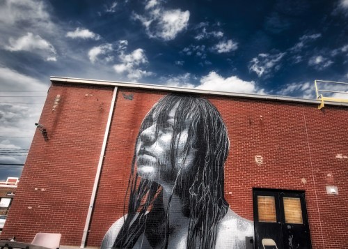 A large mural of a woman with wet hair on a brick wall, under a cloudy blue sky.