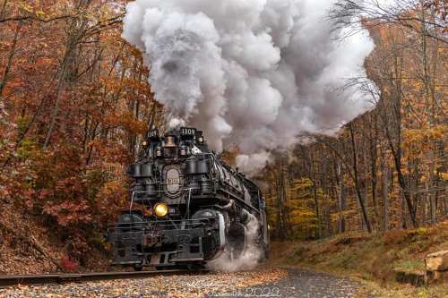 A vintage steam locomotive chugs along a forested track, billowing white smoke amidst colorful autumn foliage.
