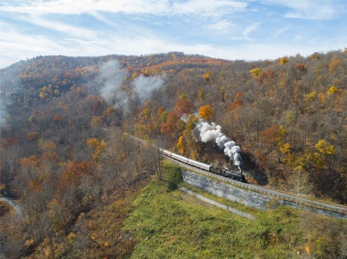 A steam train travels through a colorful autumn landscape with rolling hills and trees in vibrant fall foliage.