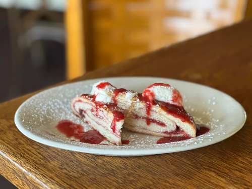 A slice of rolled cake with strawberry sauce and whipped cream, dusted with powdered sugar on a white plate.