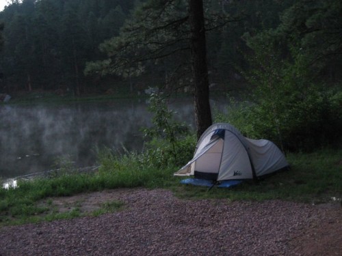 A tent by a misty lake surrounded by trees, with a serene, tranquil atmosphere in the early morning light.