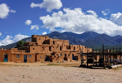 Adobe-style buildings under a blue sky with clouds, surrounded by mountains and a dirt landscape.