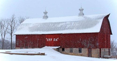 A red barn in winter, covered in snow, with the words "UFF DA" painted on its side. Trees are bare in the background.