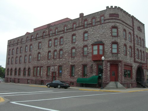 Historic stone building with red accents, featuring multiple windows and a distinctive entrance, located at a street corner.