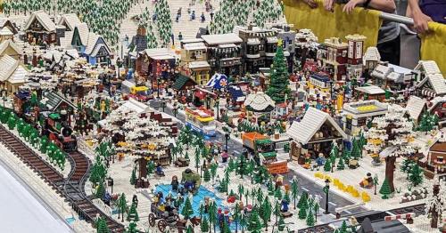 A detailed LEGO winter village scene with buildings, trees, a train track, and miniature figures.