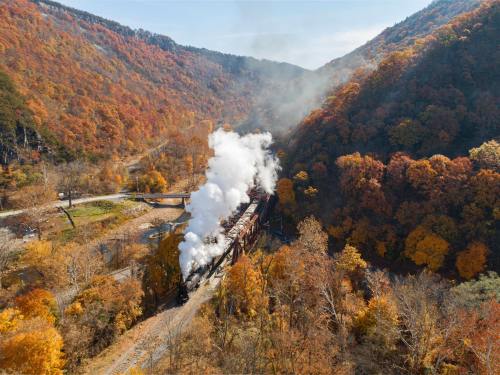 A steam train travels through a colorful autumn landscape, surrounded by mountains and vibrant fall foliage.