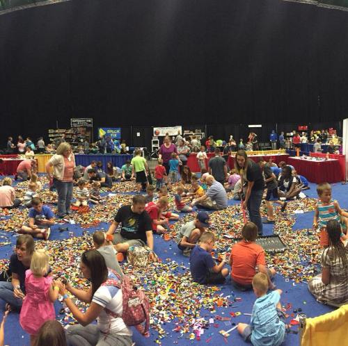 A large group of children and adults play with colorful building blocks on a blue carpet in a busy event space.