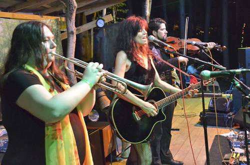 A band performs on stage with a trumpet, guitar, and violin, set in a cozy outdoor venue.