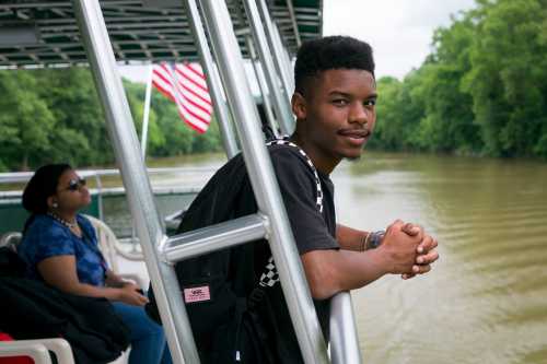 A young man leans on a railing by a river, while a woman sits nearby, both enjoying a scenic boat ride.