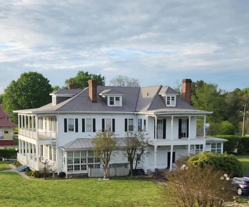 A large white two-story house with a wraparound porch, surrounded by green grass and trees under a cloudy sky.