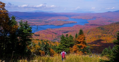 A person walks through colorful autumn foliage overlooking a lake and mountains in the distance.