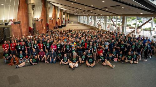 A large group of people in matching shirts poses together in a spacious indoor venue with large windows.