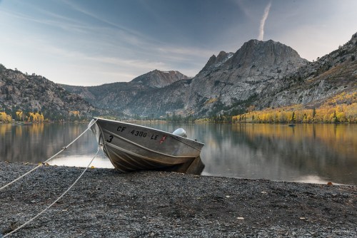 A small boat is moored on a rocky shore, with mountains and autumn trees reflected in the calm lake water.