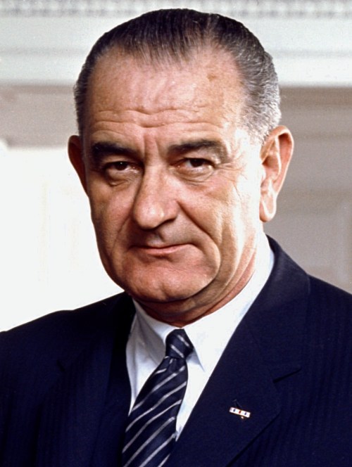 A serious-looking man in a suit, with slicked-back hair, standing against a light background.