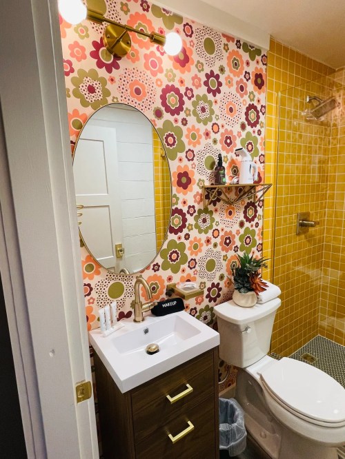 Brightly decorated bathroom with floral wallpaper, a round mirror, modern sink, and yellow tiled shower.