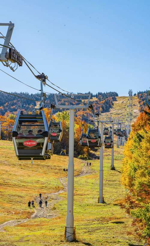 A scenic view of a gondola lift on a mountain, surrounded by autumn foliage and hikers on a trail below.