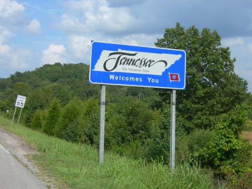 A welcome sign for Tennessee, featuring the state name and flag, with greenery and a road in the background.