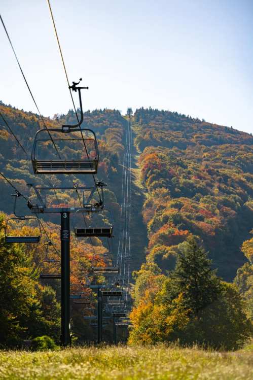 A ski lift ascends a mountain covered in vibrant autumn foliage under a clear blue sky.
