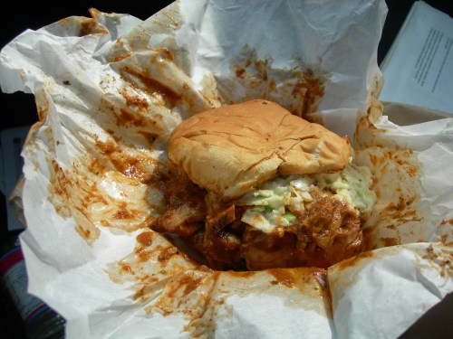 A messy sandwich with shredded meat and coleslaw, wrapped in crumpled paper, with sauce stains around it.