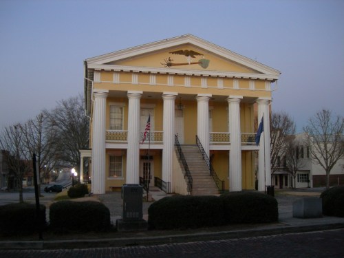 Historic building with a yellow facade, tall white columns, and stairs, set in a quiet street at dusk.