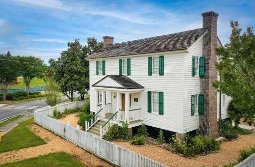 A two-story white house with green shutters, surrounded by a garden and a white picket fence, under a blue sky.