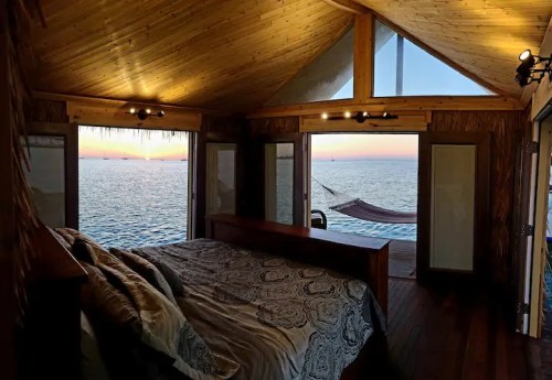 Cozy bedroom with a wooden interior, large windows overlooking a serene ocean sunset and a hammock outside.