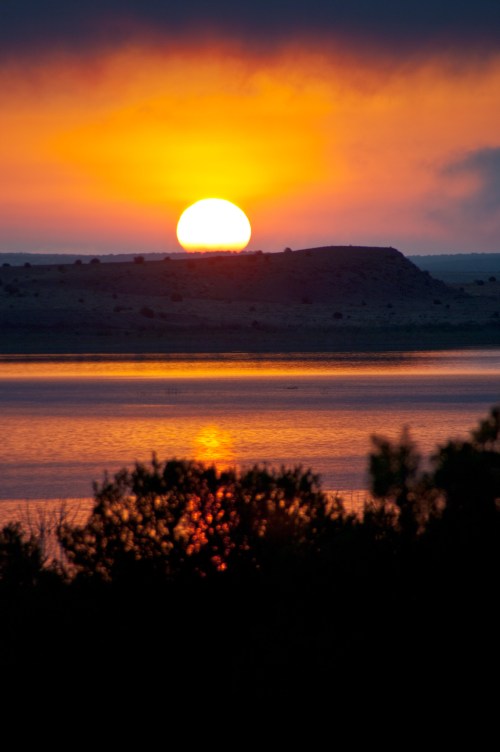 A vibrant sunset over a calm lake, with silhouettes of bushes in the foreground and hills in the background.
