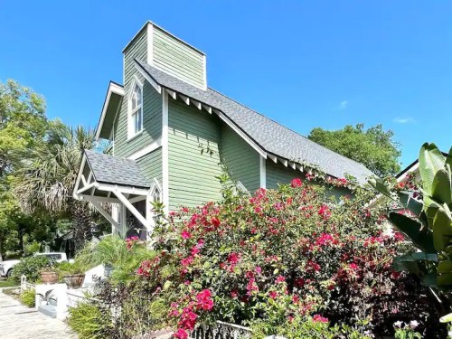 A charming green house surrounded by vibrant pink flowers and lush greenery under a clear blue sky.
